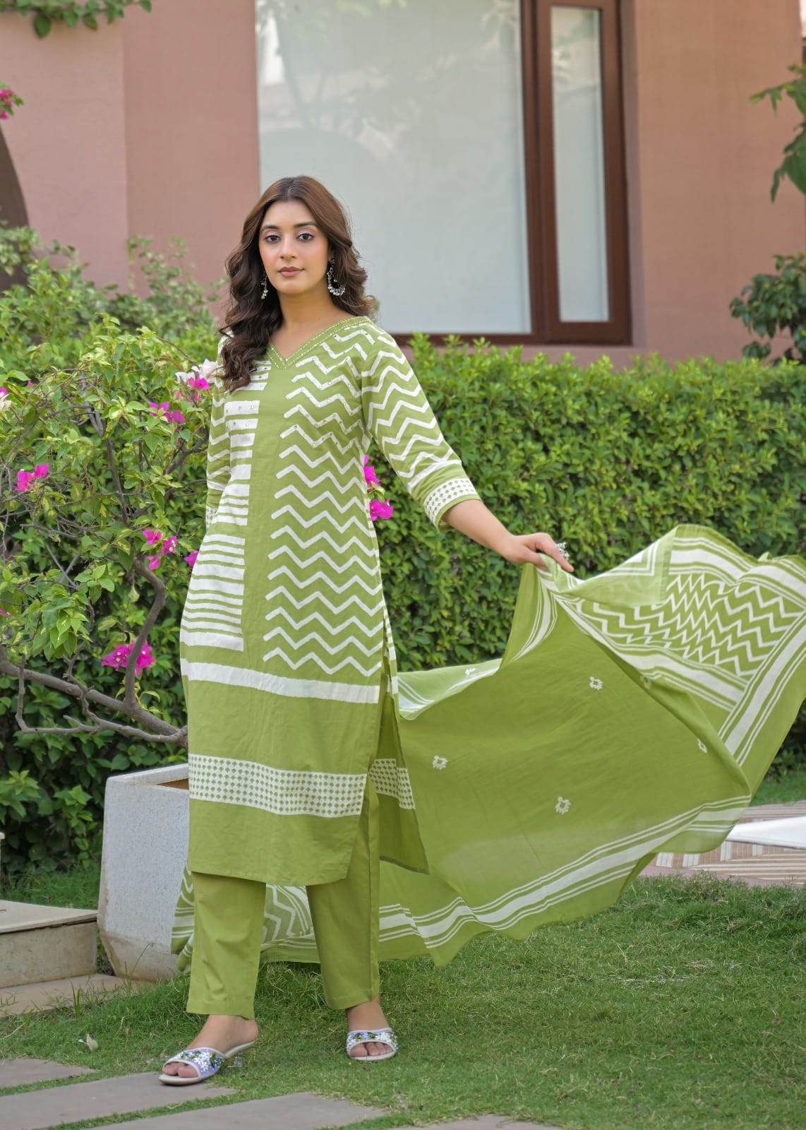 A woman stands outdoors on a lawn wearing a light green and white printed ethnic three-piece suit, holding up the matching dupatta to show its pattern.