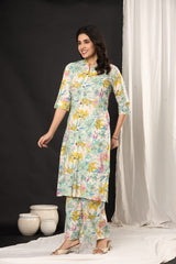 A woman modeling a light green three-piece kurta set with a colorful floral print, standing in a studio with a black backdrop and white drape.
