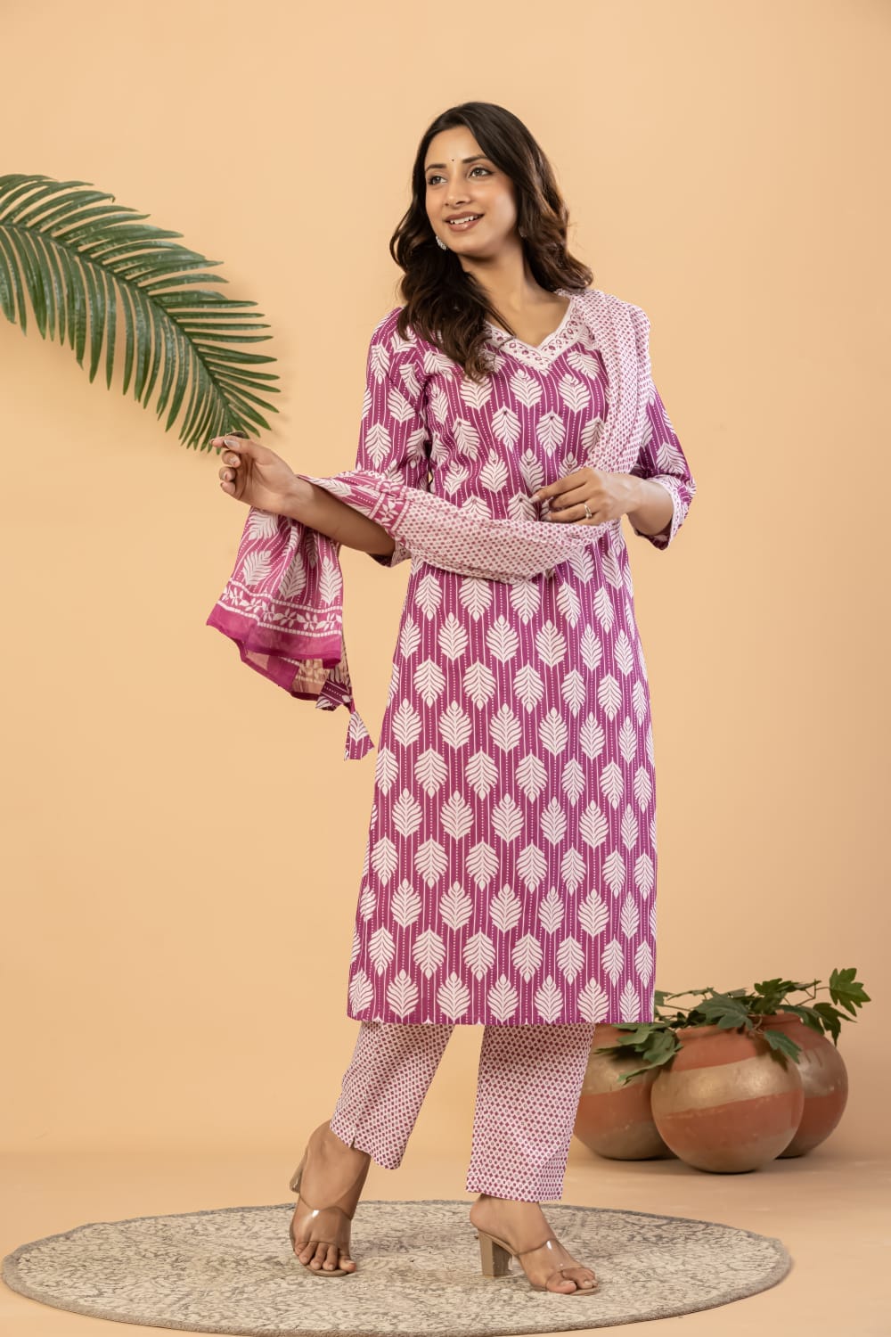A woman models a three-piece printed Indian ethnic suit consisting of a magenta kurta with white motifs, matching trousers, and a dupatta, set against a warm beige background with decorative props.