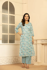 A woman modeling a light blue and white printed kurta set with matching striped pants, standing in front of a cream-colored wall with an arched window.