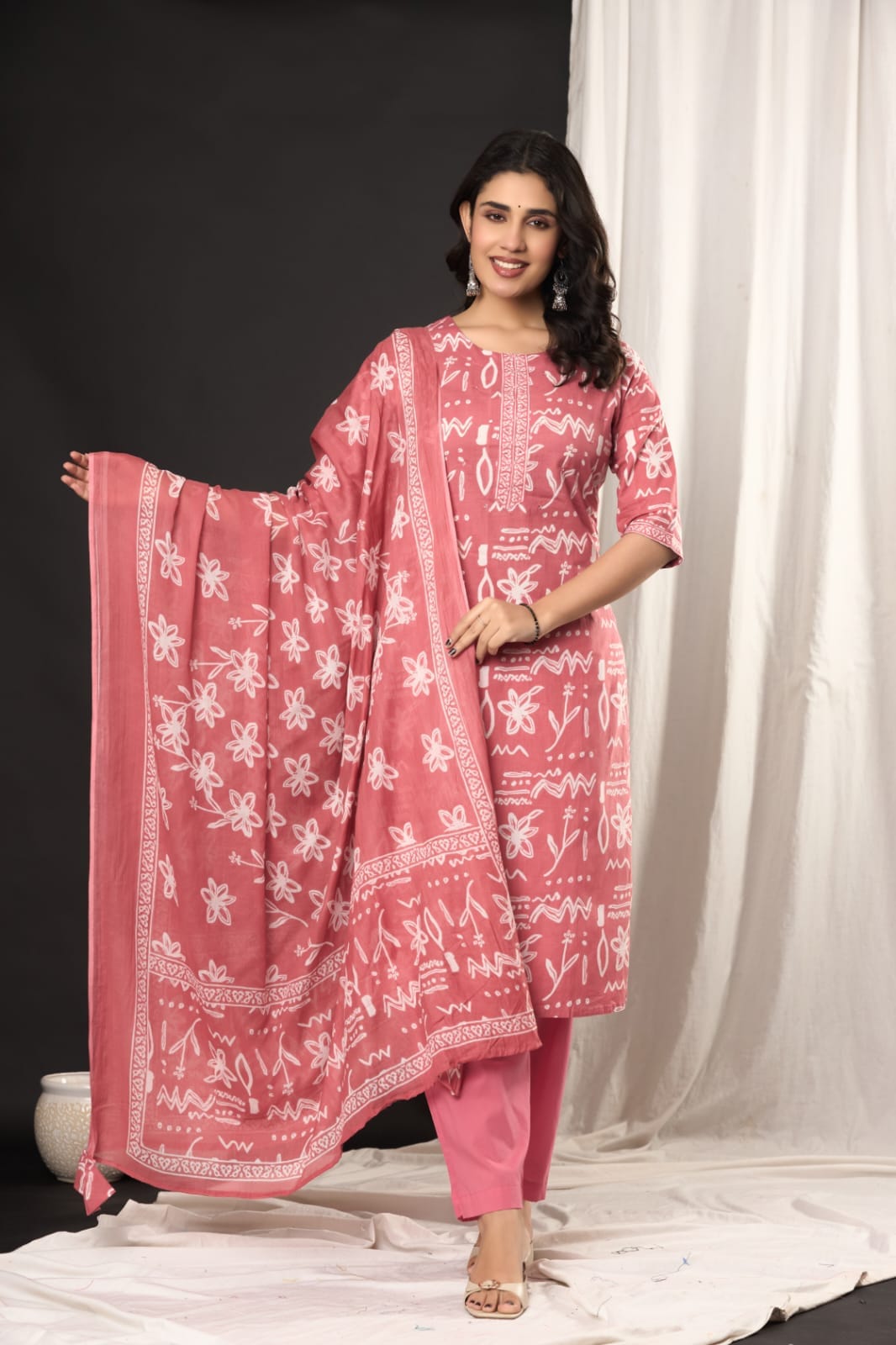 A woman models a pink three-piece salwar kameez suit with white block prints, consisting of a tunic, matching trousers, and a dupatta, set against a split black and white studio backdrop.