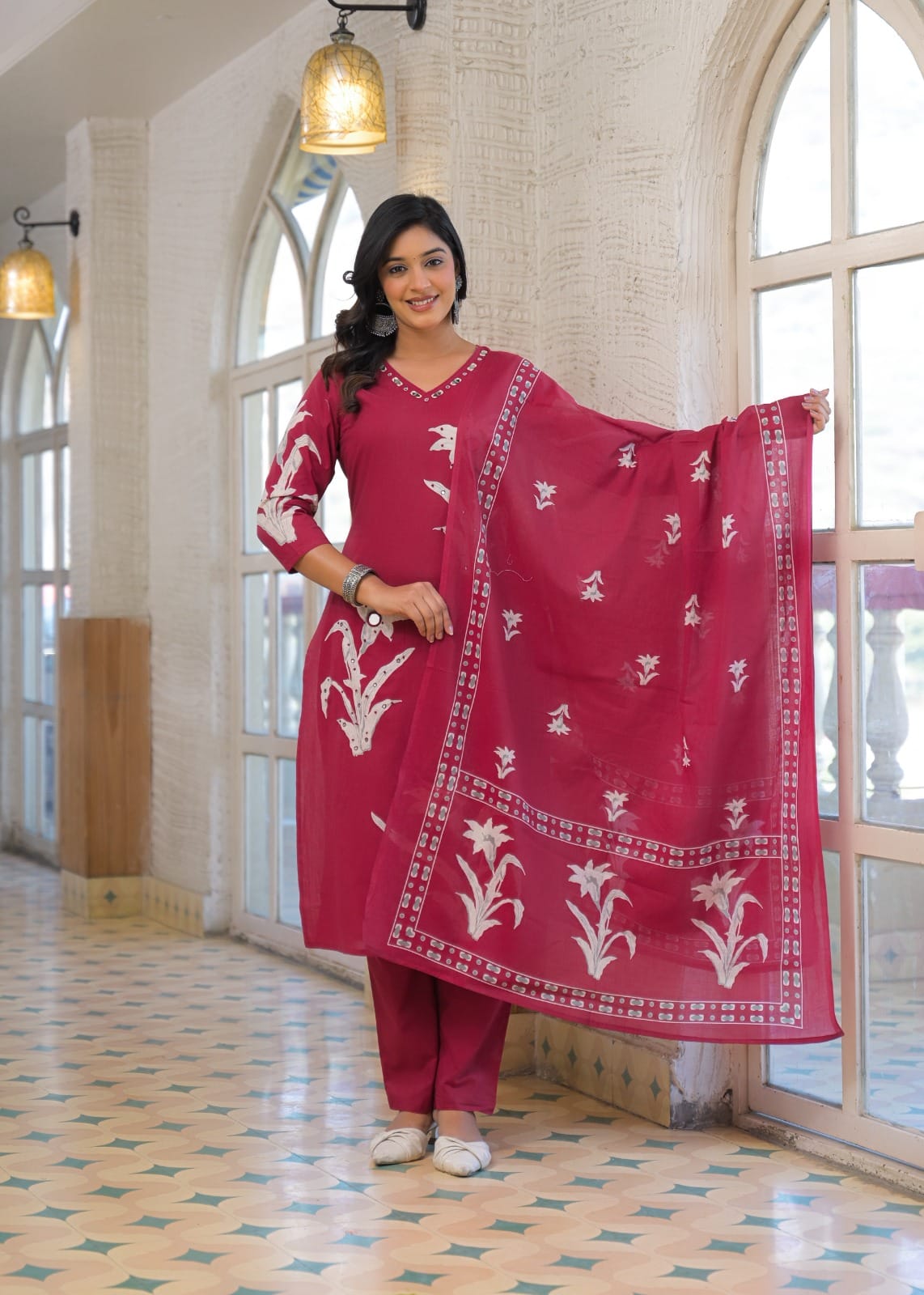 A woman models a traditional Indian salwar kameez suit in a bright indoor setting with arched windows. The set is a deep magenta pink with white floral embroidery, including a matching dupatta.
