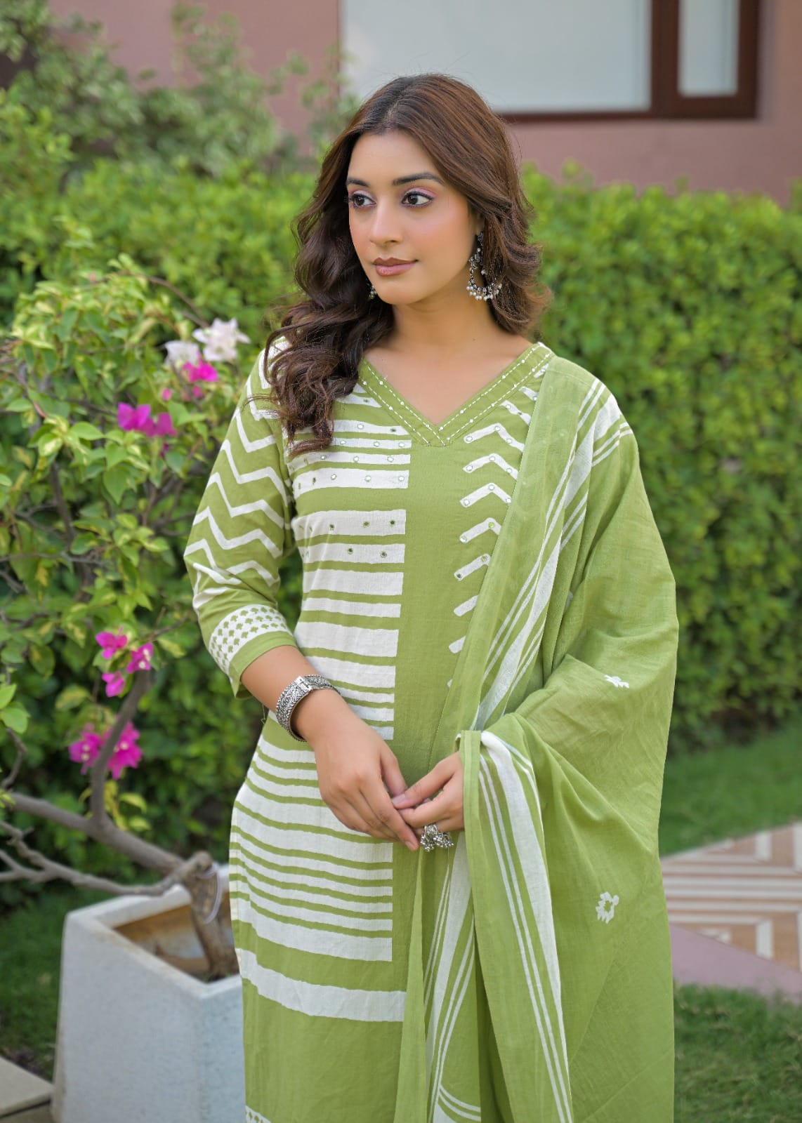 A woman wearing a light green and white printed ethnic kurta set with a matching dupatta, standing in front of a green garden hedge.