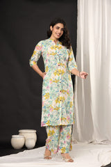A woman modeling a light-colored floral printed kurta and matching palazzo pants set, standing in a studio with a black backdrop on the left and white draped fabric on the right.