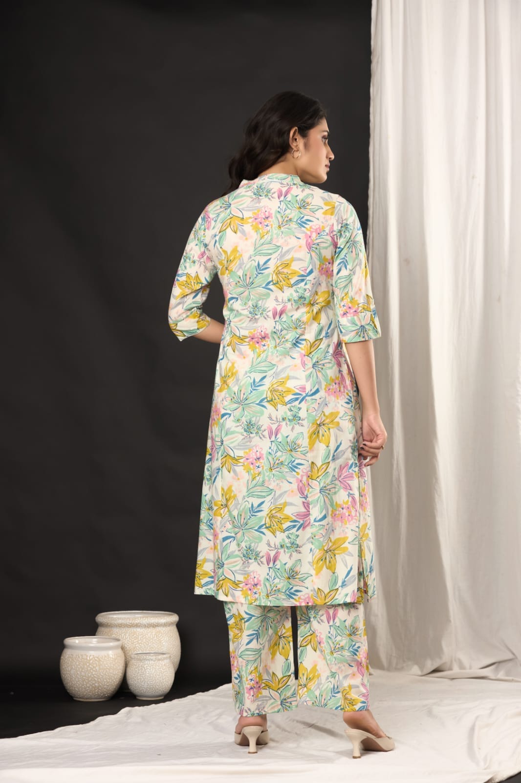 A woman stands with her back to the camera wearing a long-sleeved floral printed kurta and matching pants. The outfit features a multicolor floral pattern on a light cream base.