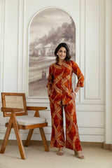 A woman models a rust-orange and beige patterned two-piece set consisting of a long-sleeve top and matching wide-leg trousers, standing in a white paneled room next to a wooden chair.