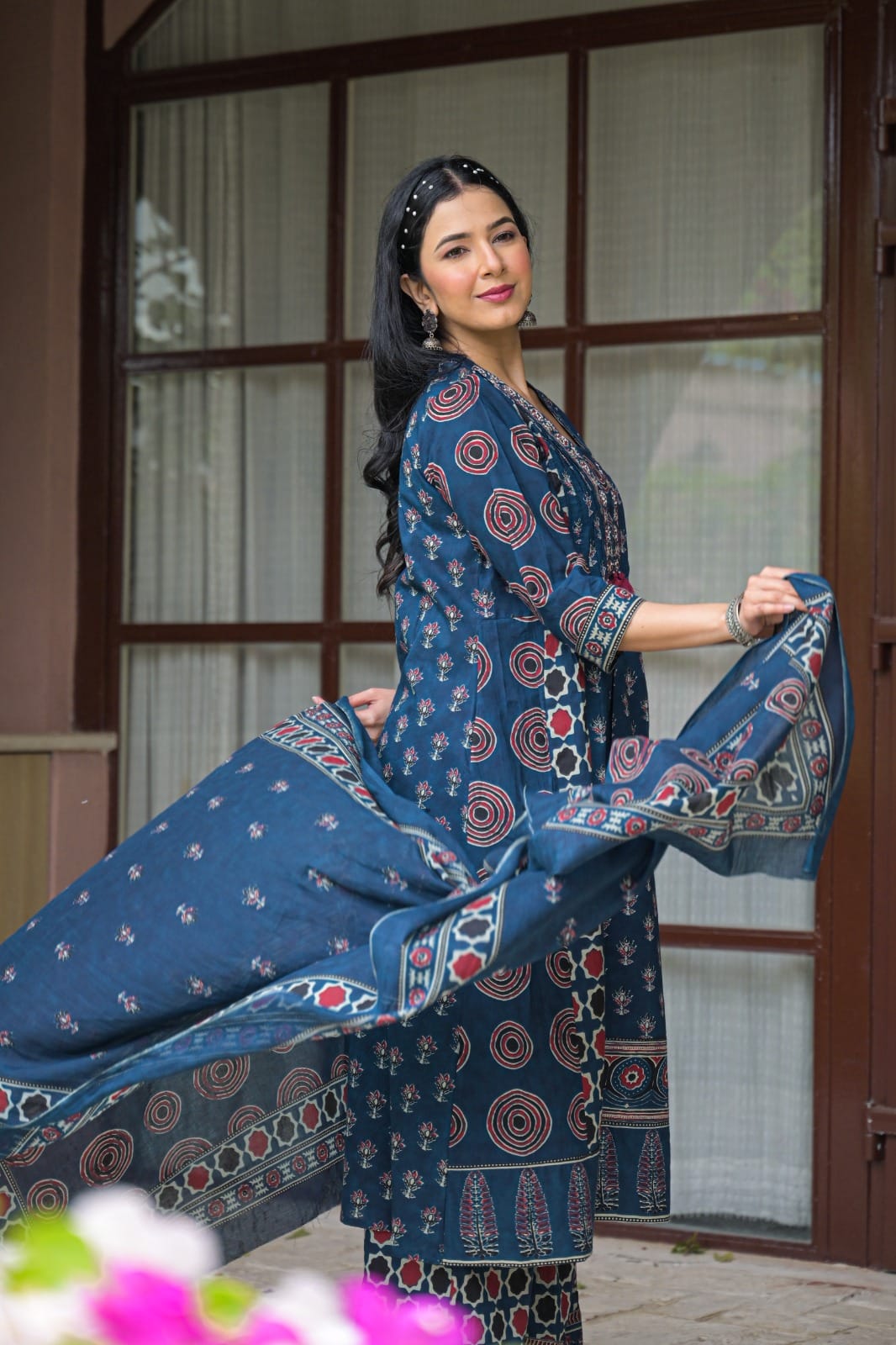 A woman modeling a navy blue ethnic salwar kameez suit with red and white block prints, holding out the matching dupatta in front of a wooden window.