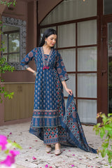 A woman models a traditional blue printed salwar kameez suit with a matching dupatta in front of a rustic building with brown wooden frames.