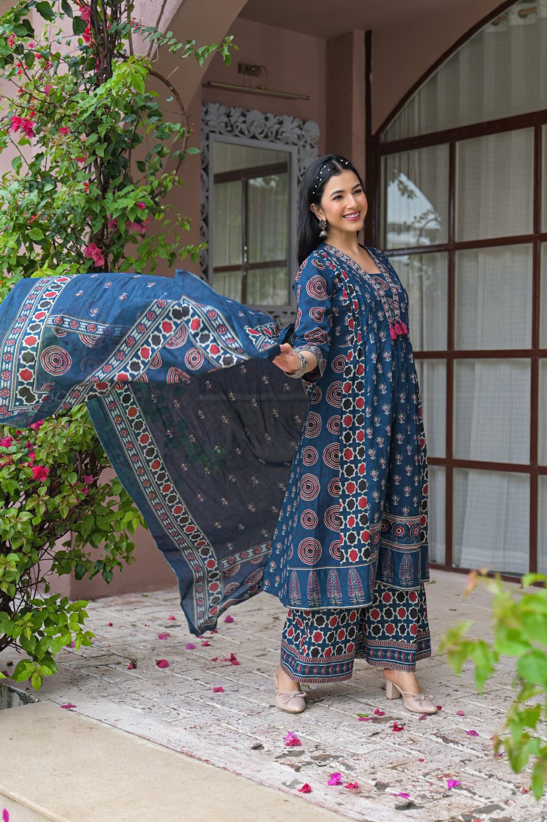A woman modeling a traditional blue printed salwar kameez suit with a matching dupatta, standing in front of a pink building with green plants.