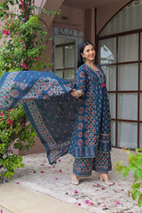 A woman modeling a traditional blue printed salwar kameez suit with a matching dupatta, standing in front of a pink building with green plants.