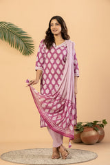 A woman models a three-piece Indian ethnic suit set featuring a magenta and white printed kurta, matching trousers, and a coordinating dupatta, set against a warm beige background with decorative plants.