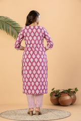 A woman stands with her back to the camera wearing a pink and white patterned kurta with matching patterned pants. The setting has a warm beige background with decorative potted plants and a palm leaf.