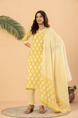 A woman models a yellow and white printed ethnic salwar kameez suit with a matching dupatta, standing on a round rug against a beige background with decorative plants.