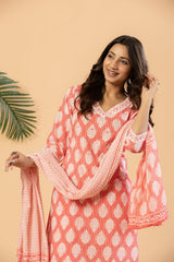 A woman modeling a coral pink and white printed kurta with matching dupatta against a warm beige background with a green palm leaf on the left.