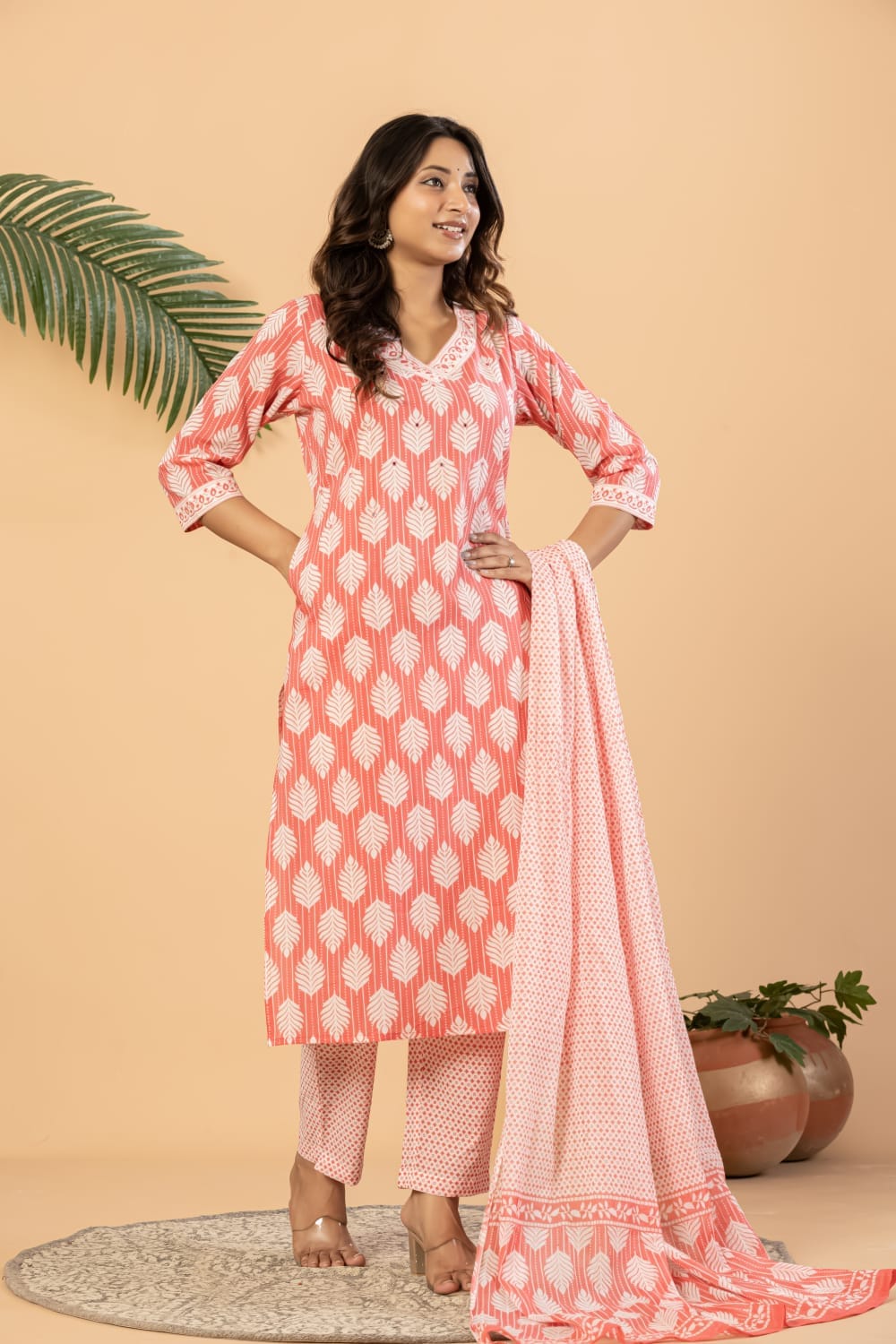 A woman models a three-piece Indian ethnic suit set consisting of a coral pink kurta with white geometric patterns, matching trousers, and a coordinating dupatta, set against a warm beige background with decorative plants.