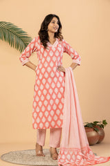 A woman models a three-piece Indian ethnic suit set consisting of a coral pink kurta with white geometric patterns, matching trousers, and a coordinating dupatta, set against a warm beige background with decorative plants.