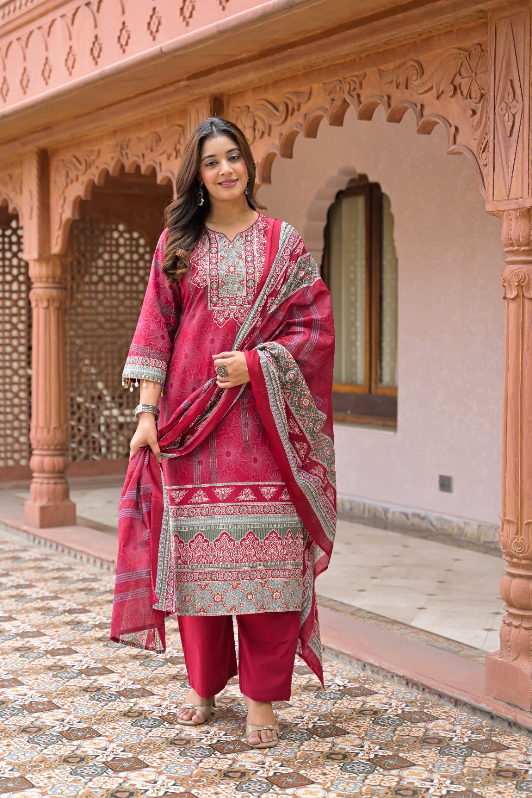 A woman models a traditional Indian salwar kameez suit set in a pink courtyard. The set includes a magenta-pink kurta with intricate multicolor embroidery, matching pink trousers, and a coordinating pink dupatta with a gray and gold border.