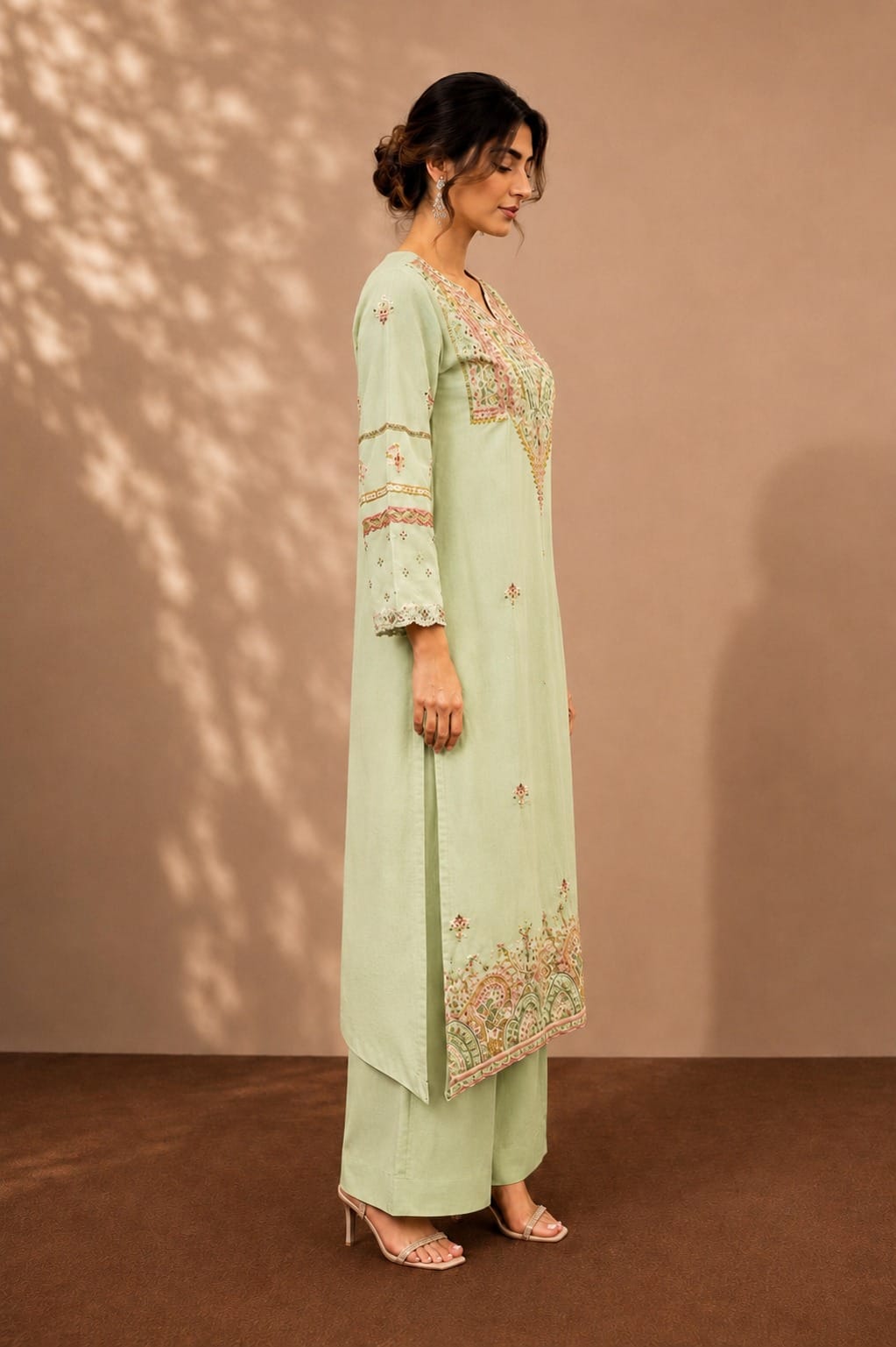A woman models a light sage green three-piece kurta set with floral embroidery, standing against a warm brown backdrop with soft leaf shadows.