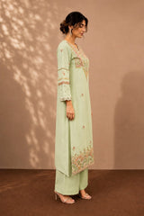 A woman models a light sage green three-piece kurta set with floral embroidery, standing against a warm brown backdrop with soft leaf shadows.