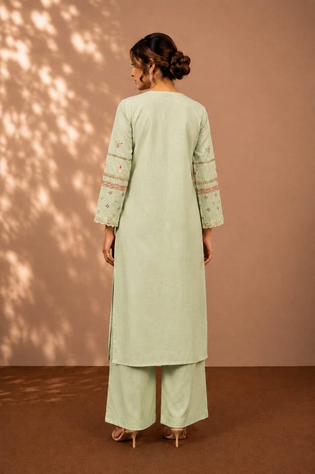 A woman stands with her back to the camera wearing a light sage green kurta and matching wide-leg pants. The kurta has embroidered details on the sleeves, and the outfit is styled with beige high-heeled sandals against a warm brown backdrop with soft leaf shadows.