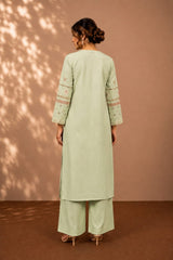 A woman stands with her back to the camera wearing a light sage green kurta and matching wide-leg pants. The kurta has embroidered details on the sleeves, and the outfit is styled with beige high-heeled sandals against a warm brown backdrop with soft leaf shadows.