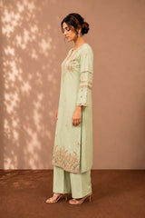 A woman models a light sage green traditional Indian outfit consisting of a long kurta with floral embroidery and matching wide-leg trousers, set against a warm brown backdrop with leaf shadow patterns.
