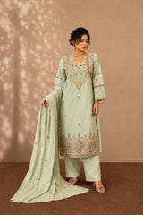 A woman models a light sage green traditional three-piece salwar kameez suit with intricate embroidery, matching trousers, and a long dupatta against a warm brown backdrop with leaf shadows.
