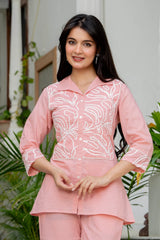 A woman modeling a light pink two-piece kurta set with white floral embroidery on the front, featuring a button-up top and matching pants, set against a soft-focus indoor background with green plants.