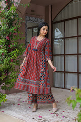 A woman wearing a red printed kurta and matching palazzo pants stands in front of a building with pink walls, arched windows, and climbing green vines.