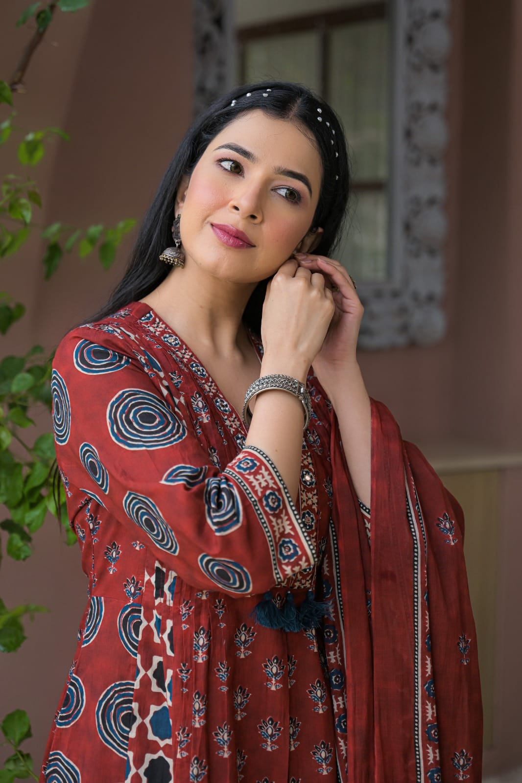 A woman wearing a rust-red printed salwar suit with blue and white motifs, featuring a matching dupatta and silver jewelry, posing in front of a warm-toned architectural background.