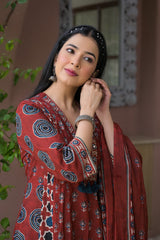 A woman wearing a rust-red printed salwar suit with blue and white motifs, featuring a matching dupatta and silver jewelry, posing in front of a warm-toned architectural background.