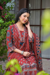 A woman wearing a rust-red printed salwar suit with blue and white motifs, seated outdoors in front of a building with green foliage in the foreground.