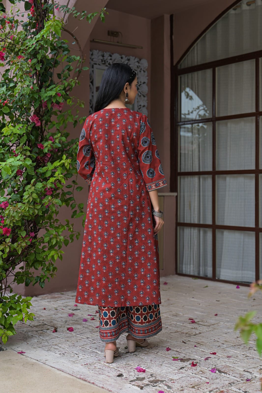 A woman viewed from behind wearing a rust-red printed kurta and matching printed pants, standing in a courtyard with a pink wall, window, and green climbing plants.