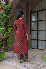 A woman viewed from behind wearing a rust-red printed kurta and matching printed pants, standing in a courtyard with a pink wall, window, and green climbing plants.