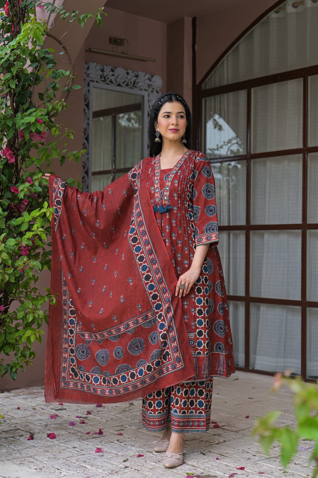 A woman models a traditional Indian salwar kameez suit set in a courtyard setting. The set is a deep reddish-brown color with intricate blue and white block prints.