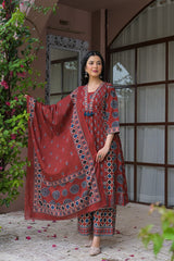 A woman models a traditional Indian salwar kameez suit set in a courtyard setting. The set is a deep reddish-brown color with intricate blue and white block prints.