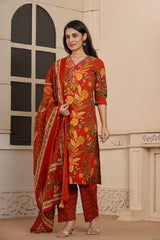 A woman models a traditional Indian salwar kameez suit set in a warm beige indoor setting. The set features a rust-red kurta with a vibrant orange, yellow, and green floral print, matching patterned trousers, and a coordinating dupatta with a gold border.