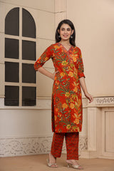 A woman modeling a traditional Indian salwar kameez set featuring a vibrant orange-red kurta with a yellow and green floral print, paired with matching patterned trousers, set against an indoor architectural backdrop.