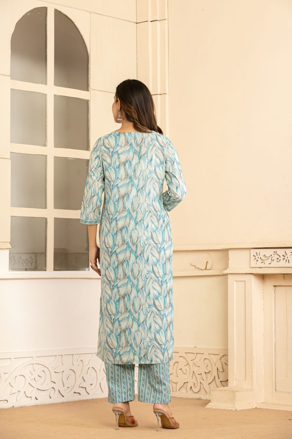 A woman stands with her back to the camera wearing a light blue and white patterned kurta with matching striped pants, standing in front of a cream-colored wall with a window.
