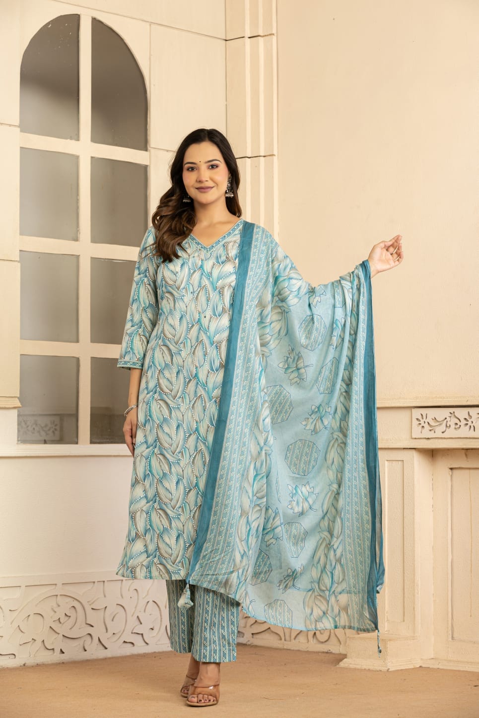 A woman models a light blue and white printed ethnic three-piece suit with a matching dupatta in an indoor setting with cream-colored walls and a window.