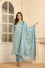 A woman models a light blue and white printed ethnic three-piece suit with a matching dupatta in an indoor setting with cream-colored walls and a window.