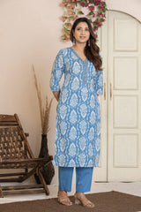 A woman modeling a light blue and white printed kurta with matching blue cropped pants, standing in a styled indoor setting with a wooden chair, dried grass in a vase, and a cream-colored door.