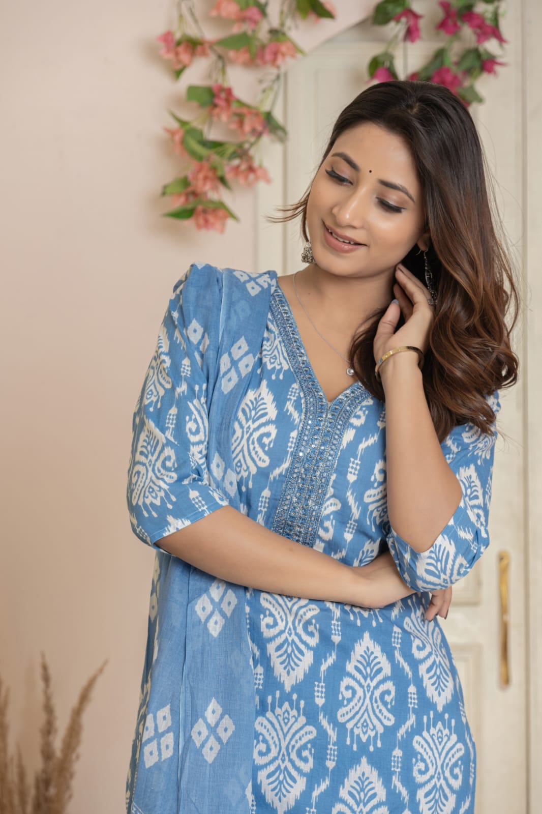 A woman wearing a light blue cotton kurta with a white ikat pattern, standing in front of a beige wall with decorative pink flowers.