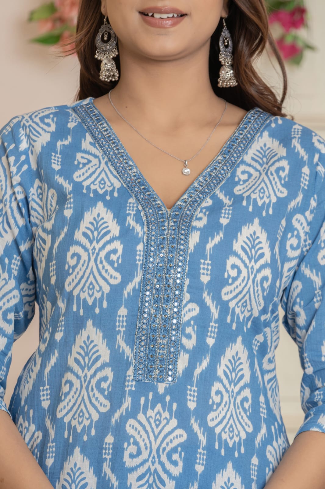 A close-up of a woman wearing a light blue and white patterned kurta with detailed silver embroidery around the V-neckline, paired with silver earrings and a necklace.