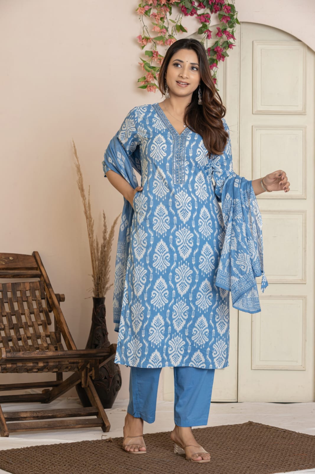 A woman modeling a light blue and white printed kurta set with matching pants and dupatta, standing in a styled indoor setting with a wooden chair and door in the background.