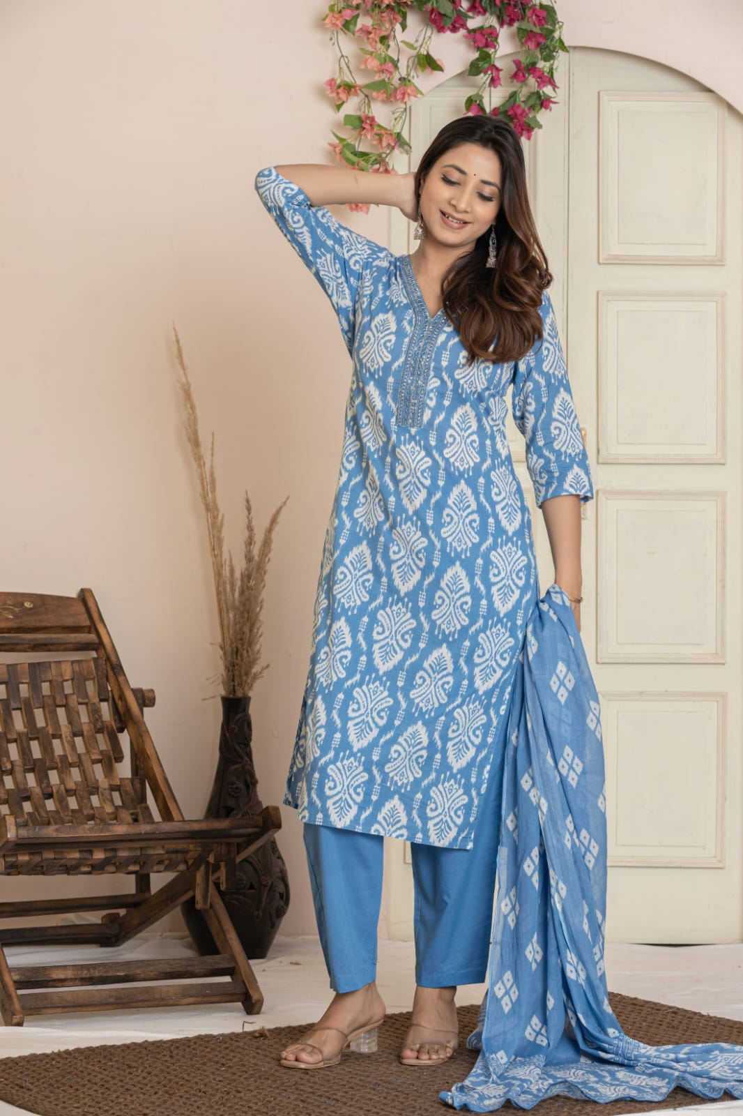 A woman models a blue and white printed Indian salwar kameez suit set, featuring a long kurta, matching trousers, and a dupatta, posed in front of a neutral beige wall with a wooden chair and door.