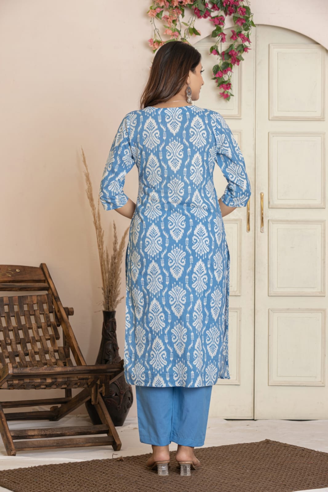 A woman stands with her back to the camera wearing a light blue and white patterned kurta with blue pants in an indoor setting with a wooden chair and cream-colored doors.