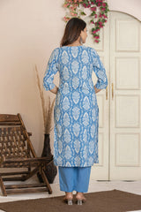 A woman stands with her back to the camera wearing a light blue and white patterned kurta with blue pants in an indoor setting with a wooden chair and cream-colored doors.