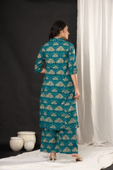 A woman stands with her back to the camera wearing a teal blue and beige printed kurta and matching palazzo pants. She is photographed in a studio setting with a black backdrop on the left, a white draped curtain on the right, and two decorative ceramic pots on the floor.
