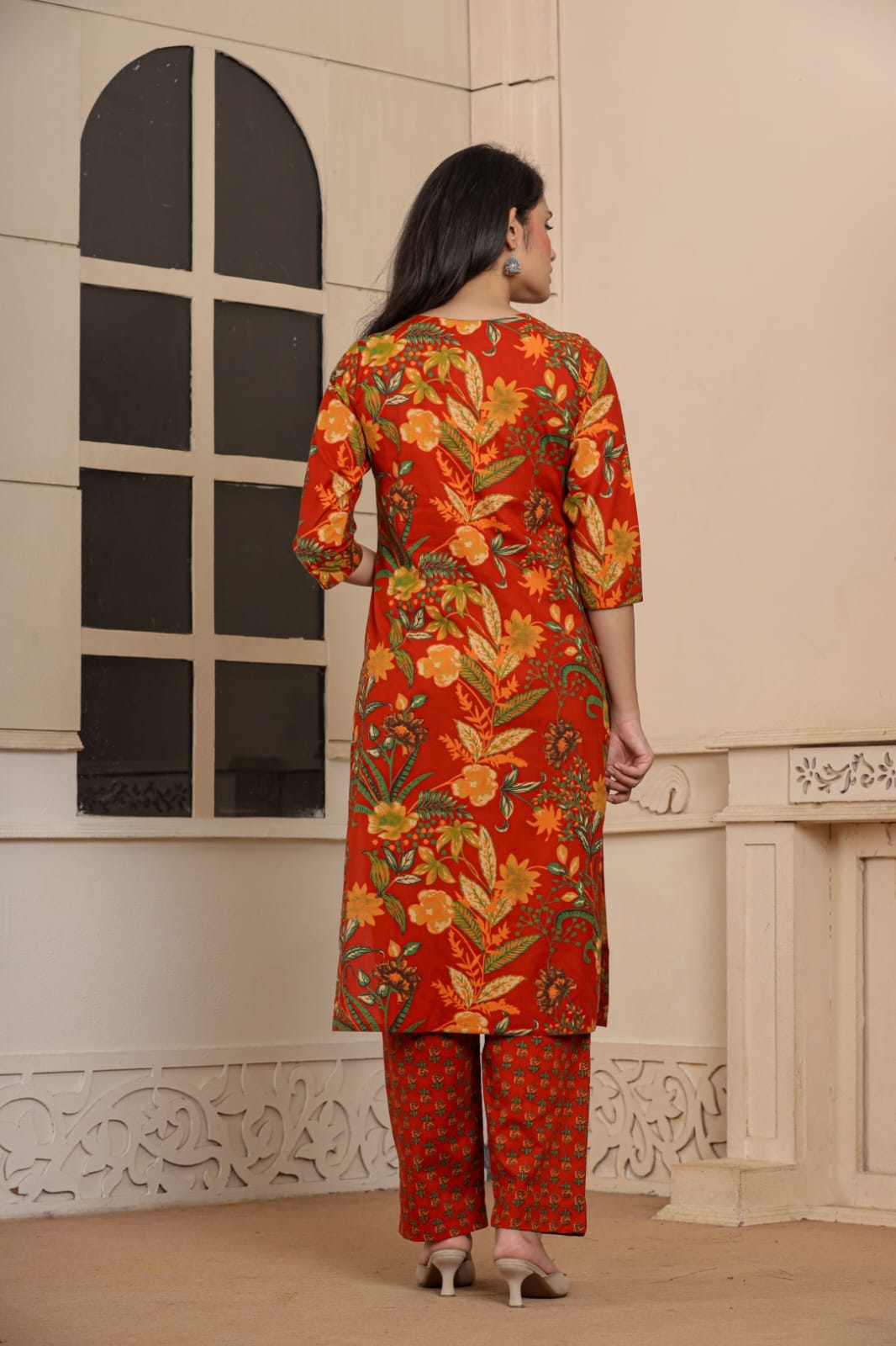 A woman viewed from behind wearing a red-orange printed kurta and matching patterned pants in an indoor setting with cream-colored walls and a decorative window.